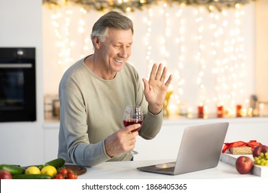 Virtual Christmas Party. Senior Man Having Video Call At Laptop Celebrating Xmas Holiday With Distant Family Online Drinking Wine In Kitchen At Home. Elderly Gentleman Toasting Waving Hand To Computer