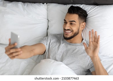 Virtual Communication. Happy Arab Guy Making Video Call With Smartphone While Relaxing In Bed At Home, Cheerful Middle Eastern Man Waving Hand At Phone Camera, Enjoying Online Meeting, Top View