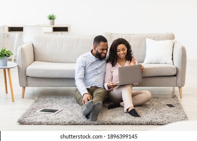 Virtual Communication. Happy black couple having online video conference with friends or family using computer, waving hand to web camera, sitting on the floor carpet in bedroom at home - Powered by Shutterstock