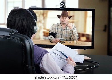 Virtual Doctor In Headset Reviews Blood Pressure Graph With On-line Patient In Monitor. Cowboy Sits At Wooden Desk, Holds A Pill By Hand Checks His Blood Pressure And Looks Directly At The Physician.