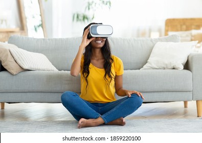 Virtual entertainments concept. Full length portrait of happy black woman in VR headset sitting on floor at home, playing interactive video games, exploring augmented reality