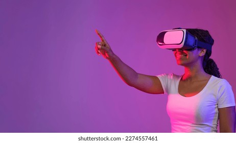 Virtual Experience. Smiling Black Woman In VR Headset Standing In Neon Light, Happy African American Female Touching Air With Hand While Playing Video Game, Enjoying Modern Technologies, Copy Space