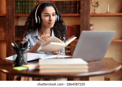 Virtual Lecture. Portrait of young lady in wireless headphones sitting at desk, holding and reading book, having online class with tutor and looking at laptop computer. Woman studying at home
