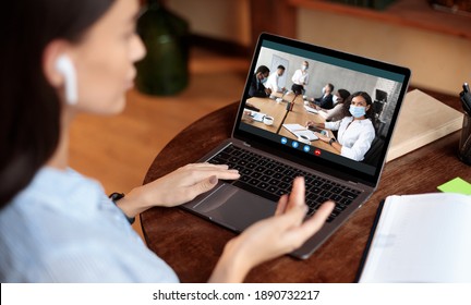 Virtual Meeting Concept. Back over the shoulder view of young woman in wireless earbuds sitting at table, using laptop having online videocall conference with colleagues wearing face masks at office