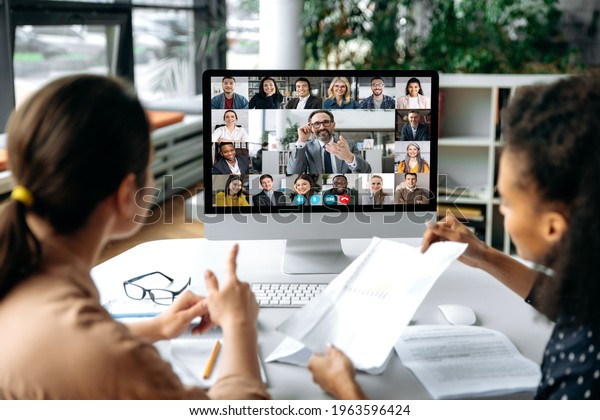 Virtual meeting online, video call. View over\
shoulders of two women to a computer screen with business leader\
and successful team, chatting by a video conference, discuss\
working issues,\
strategy