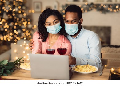 Virtual Party. African American Man And Woman In Medical Face Masks Making Video Conference With Parents Or Friends On Laptop, Sitting At Table, Holding Glasses Of Red Wine. Online Meeting At Pandemic