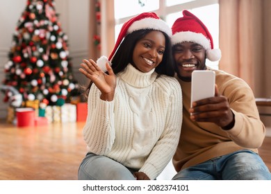 Virtual Party. Portrait of happy black couple in red santa claus hat using mobile phone, making online video conference waving hand to webcam sitting on floor in decorated living room with Xmas tree - Powered by Shutterstock