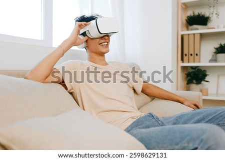 Virtual reality experience, young man smiling while wearing VR headset at home, light colored living space with a cozy sofa and natural light streaming in through windows