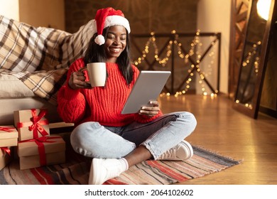 Virtual Xmas celebration. Positive black woman in Santa hat drinking coffee, sitting on floor in living room, making video call with family using tablet pc, looking at screen, shopping online - Powered by Shutterstock