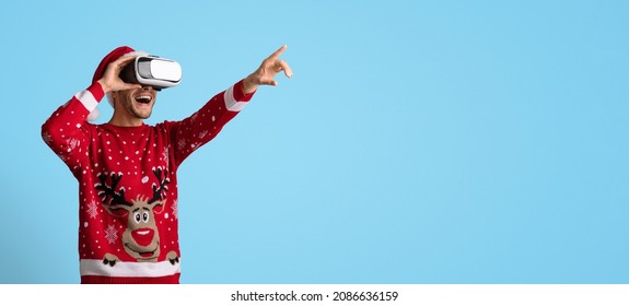 Virtual Xmas Party. Excited Young Man In Vr Glasses And Christmas Sweater Pointing Away At Copy Space Over Blue Studio Background, Cheerful Guy In Santa Hat And Goggles Enjoying Cyber Event, Panorama