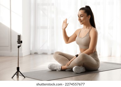 Virtual yoga classes concept. Yoga instructor millennial beautiful well-fit woman in tight sportswear sitting on fitness mat, waving at smartphone camera, greeting students, copy space - Powered by Shutterstock