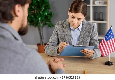 Visa interview at US embassy office with officer reviewing applicant documents, writing notes with pen. Paperwork and immigration history review by lawyer sitting at desk with United States flag