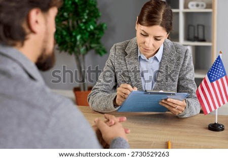 Visa interview at US embassy office with officer reviewing applicant documents, writing notes with pen. Paperwork and immigration history review by lawyer sitting at desk with United States flag