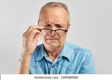 Vision Problems. An Elderly Man Takes Off His Glasses. Not Satisfied. Senior Citizen On A White Background In A Blue Shirt. Optics