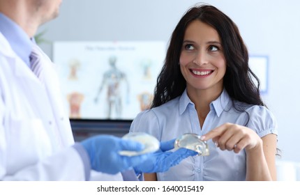 Visit To Female Breast Plastic Surgery Clinic. Young Women Are In Doctor Office At Plastic Surgery Clinic And Smiling. Doctor Shows Patient Breast Implants. Consultation With An Experienced Surgeon.