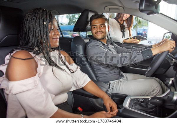 Visiting a car dealership choosing and buying a\
new car. A young emotional African-American married couple\
communicate in the car interior against the background of a car\
sales agent. Selective\
focus.