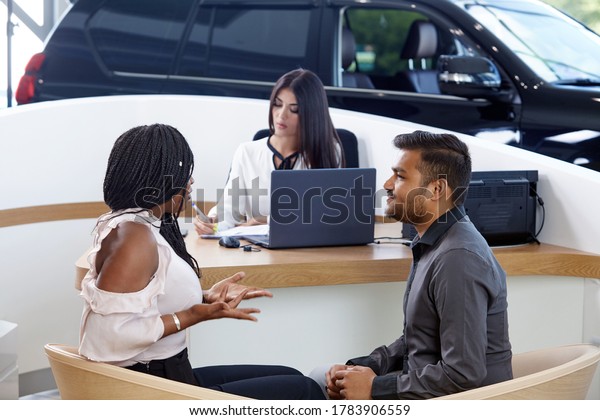 Visiting a car\
dealership choosing and buying a new car. A young emotional\
African-American married couple communicate against the background\
of a car sales agent. Selective\
focus.