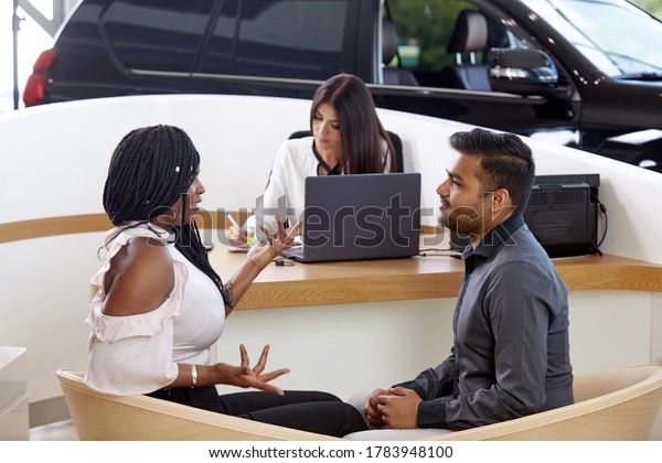 Visiting a car\
dealership choosing and buying a new car. A young emotional\
African-American married couple communicate against the background\
of a car sales agent. Selective\
focus.
