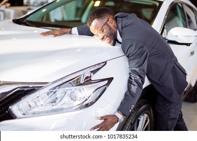 Visiting Car Dealership. Handsome Black Man Is Hugging His New Car And Smiling