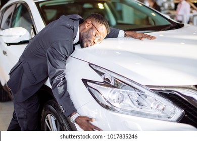 Visiting Car Dealership. Handsome Black Man Is Hugging His New Car And Smiling