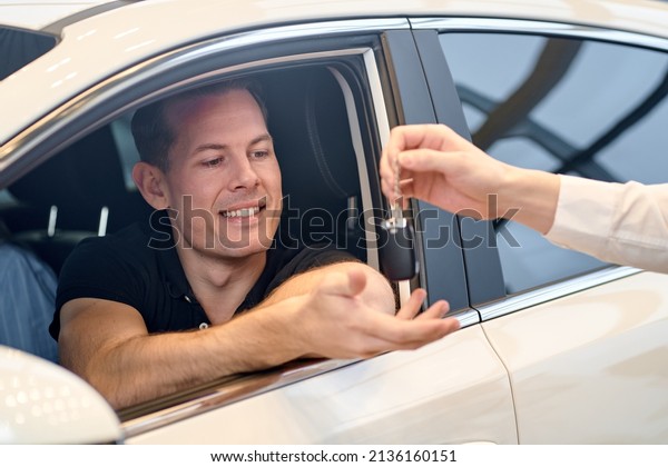 Visiting car\
dealership. positive guy is getting a car key and smiling while\
sitting in new car. young happy male getting keys by cropped male\
manager car dealer, in modern cars\
showroom
