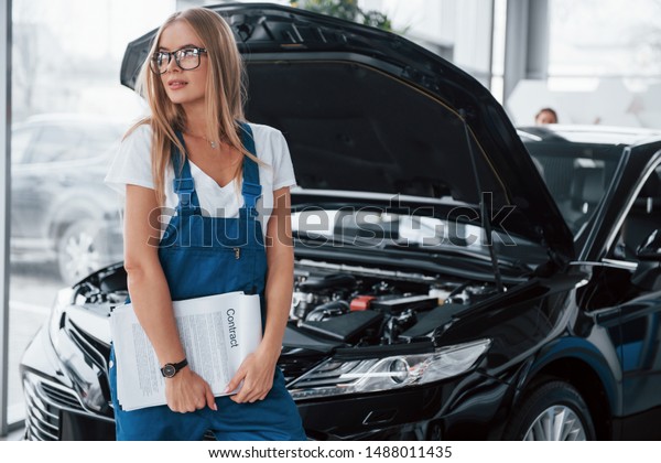 Visitor stands behind the car. Manager with notepad,\
in the white shirt and blue uniform stands against automobile with\
hood up.