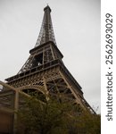 Visitors admire the grandeur of the Eiffel Tower in Paris, capturing its intricate iron lattice design. The shot is taken from a low angle, emphasizing its towering presence against a cloudy sky.