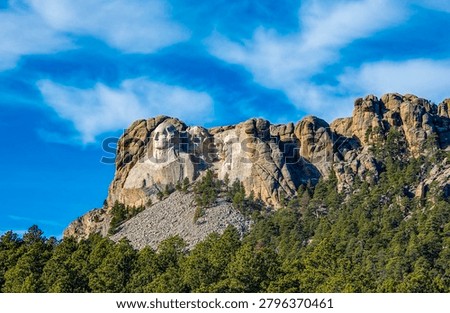 Visitors can view large stone faces of U.S. presidents carved on a mountain. The site is surrounded by trees and the sky is clear with some clouds.