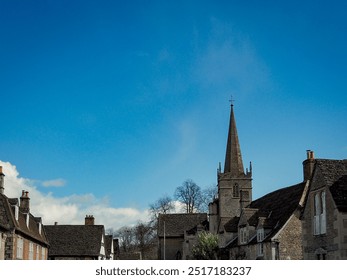 Visitors stroll through the historic streets of Lacock village on a sunny day, surrounded by stone houses and a church, with parked cars lining the road. - Powered by Shutterstock