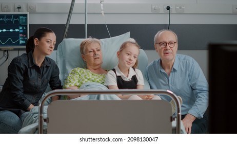Visitors watching television with elder patient in hospital ward bed. Mother and little girl visiting pensioner with sickness, looking at tv and enjoying visit with family. People supporting woman