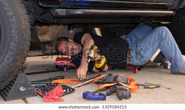 Vista, CA / USA - March 3, 2019: Older man\
laying under his truck while working on it reaches out to select a\
new tool                             \
