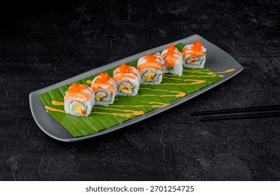 A visually appealing array of sushi rolls, elegantly presented on a gray rectangular plate lined with a fresh green banana leaf. 