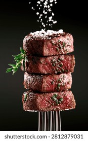 A visually appealing stack of four perfectly grilled beef steaks, seasoned with fresh herbs and coarse salt, presented on a fork against a dark background.