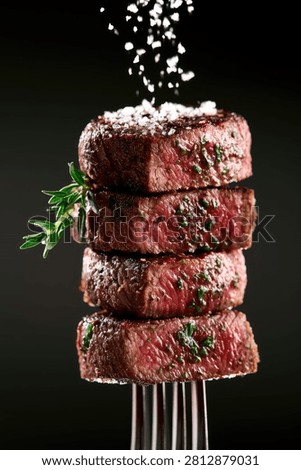 A visually appealing stack of four perfectly grilled beef steaks, seasoned with fresh herbs and coarse salt, presented on a fork against a dark background.