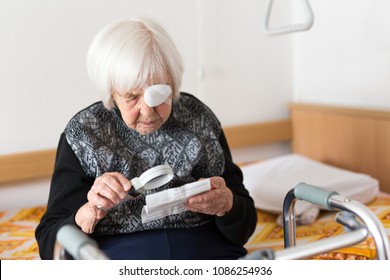 Visually Impaired Elderly 95 Years Old Woman Sitting At The Bad Trying To Read Her Medical Therapy Prescription With Magnifying Glass Due To Eyesight Problems.