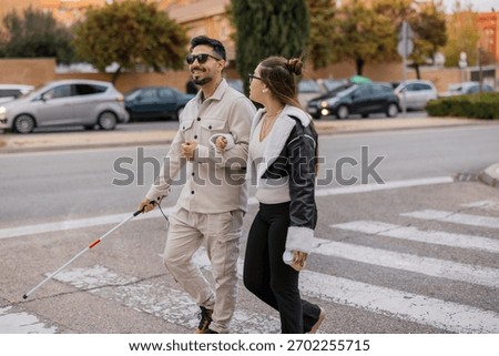 Visually impaired man using a white cane and receiving assistance from a young woman, walking across a pedestrian crosswalk in an urban environment with traffic in the background