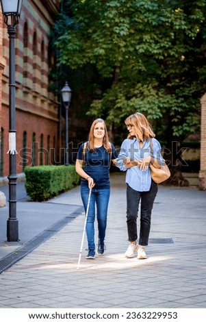 Visually impaired woman walking with female friend on the city street. Mature caucasian woman assisting her friend who is visually impaired. Full length. 