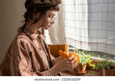 vitamin C deficiency. Millennial hipster girl peeling citrus fruit while standing by window in kitchen at home, eating oranges to boost immunity during seasonal changes. Mood boosting food - Powered by Shutterstock