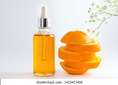 Vitamin C Serum In Cosmetic Bottle With Dropper, Sliced Orange And Flowers On White Background. Organic SPA Cosmetics With Herbal Ingredients.