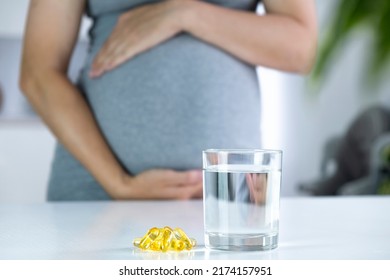 Vitamin D capsules and glass of water and pregnant woman at home interior. Healthy fatty acids nutritional supplement for prenatal support. Omega, DHA, vitamin D, fish oil for healthy pregnancy.