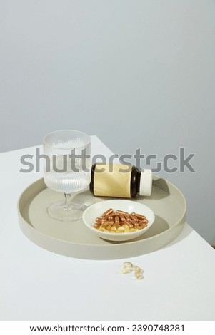 Vitamin E and hard capsules are contained in a ceramic plate, a glass of water and an unlabeled medicine bottle on a minimalist background. Mockup for dietary supplement advertising.
