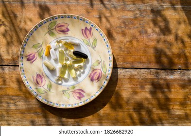 Vitamin Tablets On A Tea Plate With Floral Decorations. Supplement Capsules On An Old Wooden Table In Beautiful Dappled Light Photographed From Above