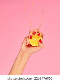 Vitamines. Female hand with bright manicure squeezes half of orange over pink background. Pop art food photography. Drops of juice fly up. Concept of healthy eating, art, fashion, creativity and ad