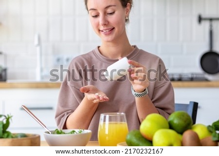 Vitamins, nutritional supplements, healthy lifestyle concept. Young woman eating breakfast and taking vitamin sitting at home in the kitchen, smiling friendly