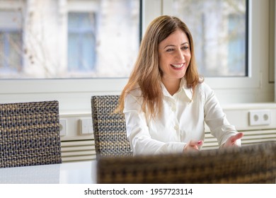 Vivacious animated woman gesturing with her hand and laughing as she sits at a conference table in a high key office or apartment with window backdrop