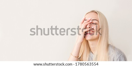 Vivacious woman giglling with her eyes screwed up in a moment of fun as she sitting against beige studio wall. Happy natural laughing young casual female covering mouth. Human face expressions