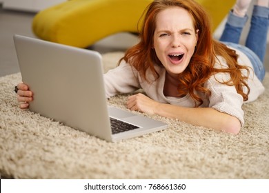 Vivacious Young Woman Laughing At The Camera With A Whimsical Expression As She Lies On A Carpet Using A Laptop