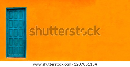 Vivid bright orange wall house facade with blue green closed door in left of large empty orange wide wall texture background space in panorama banner format with large copy space.