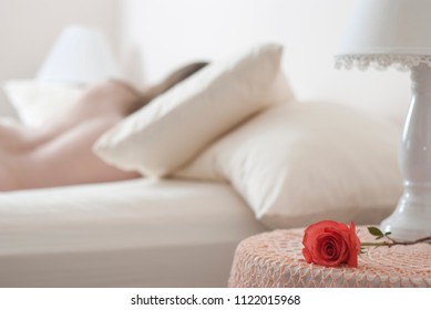 A Vivid Coral Rose Lies On A Night Stand Table In A Bedroom, With The Very Blurred And Soft Focus Image Of A Woman Sleeping In The Background