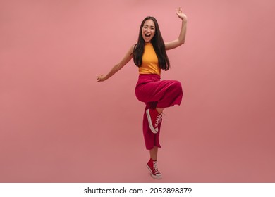 Vivid full-length image of young Asian woman in casual clothes having fun with camera. Bending leg and holding hands at sides, in crimson pants, orange T-shirt and red envelopes.